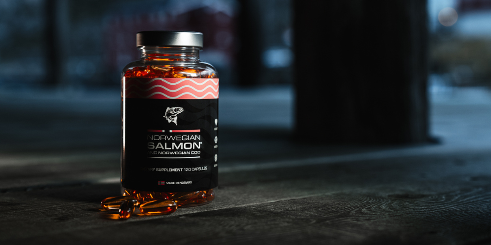 Flasche mit norwegischen Lachs- und Dorschöl-Kapseln auf einem Holzboden, beleuchtet von der Seite, mit dem Etikett 'Norwegian Salmon and Norwegian Cod – Made in Norway'.